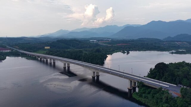 A long concrete bridge spans a calm river surrounded by dense green forest with mountains and cloudy sky in the distance at Tasik Raban