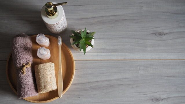Luxury eco-friendly bathroom setup with a bamboo toothbrush, natural loofah, and rolled pink towel on a wooden tray, paired with a white marble ceramic soap dispenser. Side view on a light gray wood 