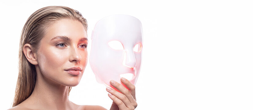 A blonde woman wearing an LED facial mask against a white background with space for text. Professional skin care, rejuvenation, innovative cosmetic technologies, skin healing, and beauty device