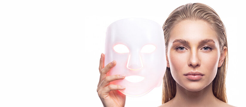 A blonde woman wearing an LED facial mask against a white background with space for text. Professional skin care, rejuvenation, innovative cosmetic technologies, skin healing, and beauty device