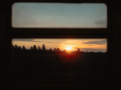 Golden sunset over a dark forest and field silhouette, framed by a retro train or bus window during a evening trip.