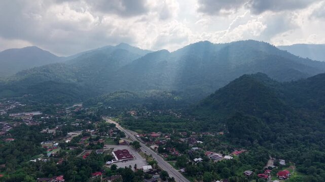 Scenic mountain range with green valley and village road under dramatic clouds and rays of sunlight creating a peaceful and natural outdoor landscape at Lenggong
