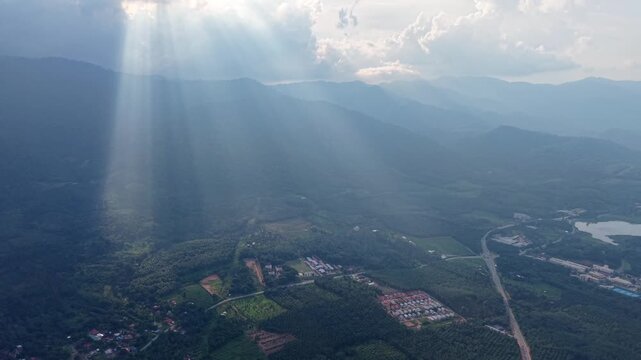 Aerial image capturing a lush green valley surrounded by mountains with sun rays piercing through dramatic clouds at Lenggong, Perak