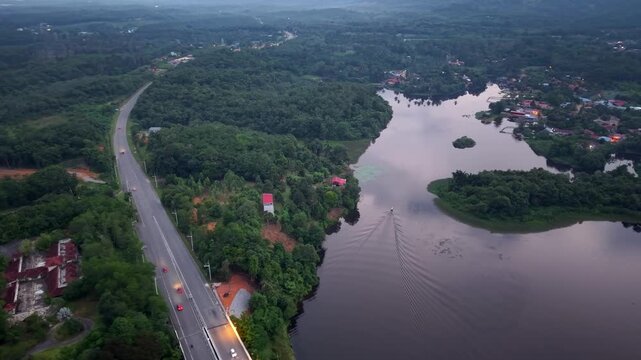 Aerial view shows a road winding through dense forests beside a calm lake. Cars travel smoothly at Tasik Raban