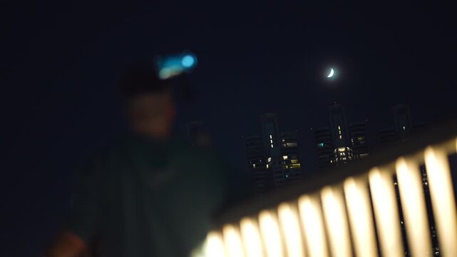 Person photographing moon eclipse with smartphone on tripod at night city skyline, man capturing lunar eclipse with mobile device and urban lights background 4k