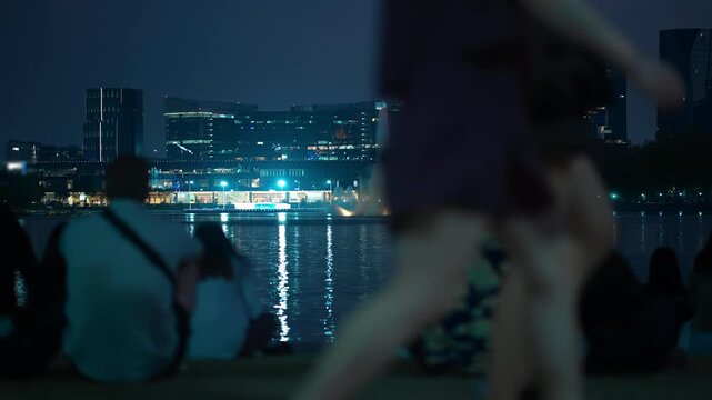 People relaxing by lake with illuminated fountain and city skyline in Benchakitti park, Night urban park scene with crowd by water and glowing fountain reflections by QSNCC Bangkok