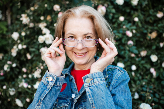 Smiling senior woman 55-60 year old with gray hair and freckles wearing eye glasses over rose flowers outdoors close up. Looking at camera.