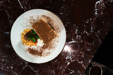 Decadent Dessert Delight: A slice of rich tiramisu, dusted with cocoa powder and adorned with a swirl of cream and mint, sits elegantly on a white plate atop a dark, marbled surface. © andreas