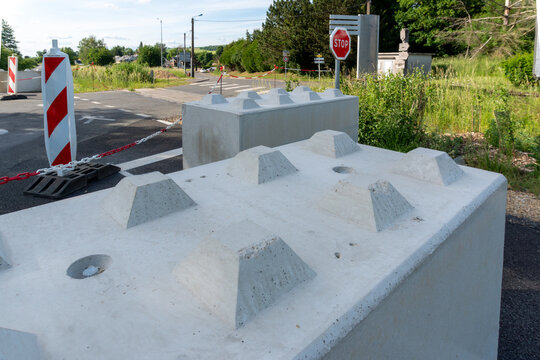 Large precast concrete blocks, striped traffic markers, plastic chains and stop signs prevent vehicles from entering a rural road. The temporary security installation represents controlled access, pub