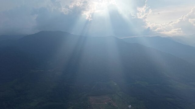 Sunlight beams pierce clouds above mountain peaks. Aerial view reveals misty slopes at Lenggong, Perak