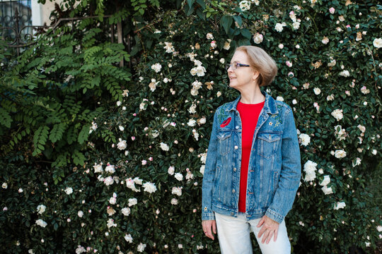 Stylish senior woman 50-55 year old wearing denim jacket and eye glasses over rose flowers outdoors.