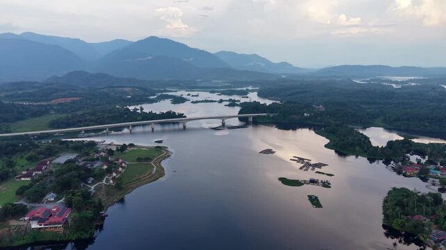 A tranquil aerial view of a wide river with a bridge crossing, surrounded by green hills and distant mountains under a cloudy sky at daylight at Tasik Raban