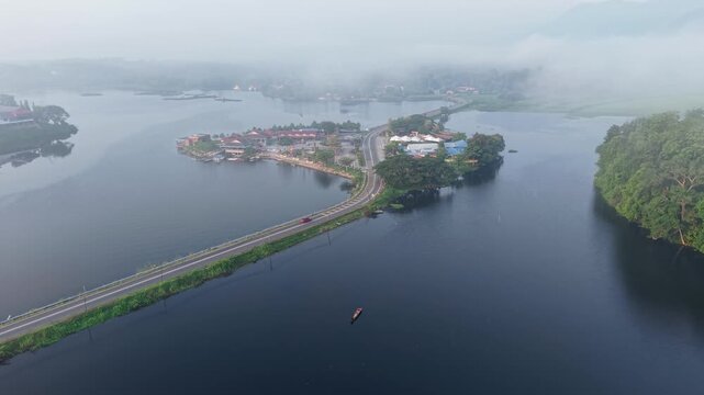 Fog blankets a serene river landscape with a narrow bridge connecting a lush green island to the mainland at Tasik Raban