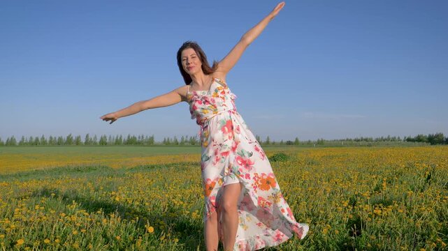 Smiling woman in flowing floral dress happily spinning and dancing in vibrant spring meadow full of yellow dandelions under clear blue sky on sunny day. Female raised arms to simulating flight