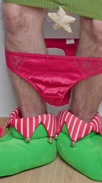 Ridiculous man wearing an elf costume and women's red underwear dancing clumsily next to christmas gift boxes on the floor in a bizarre and humorous holiday celebration scene