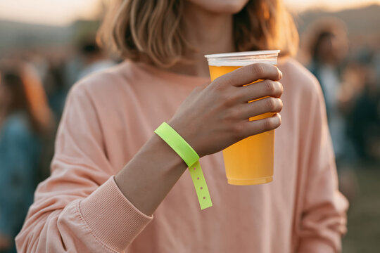 Close up of a young woman holding a plastic cup of beer at an outdoor music festival with a blank wristband for mock up template