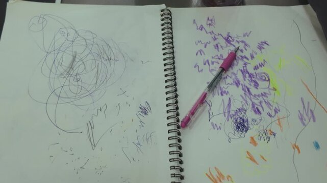 A colorful sketchbook showcases progressive child drawings with purple and orange crayon strokes while toys rest nearby on the desk surface.