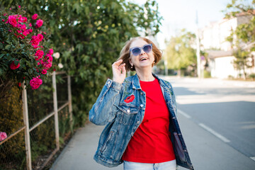 Fototapeta premium Laughing mature woman 55-60 year old wearing sun glasses and denim jacket outdoors over flowers close up. Looking at camera.