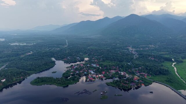Aerial green forest landscape with a calm lake, small village, winding river, and mountain range under a partly cloudy sky at daylight at Tasik Raban