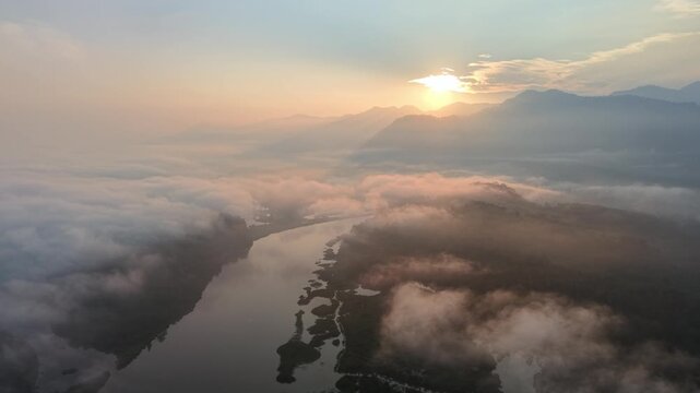A serene aerial view of a river winding through fog with a glowing sun rising behind mountains at Tasik Raban