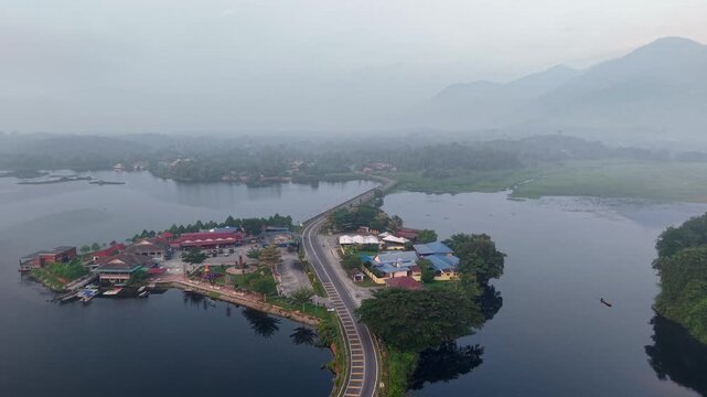 Aerial view of misty lake with island and bridge. Red roofed buildings line the shore at Tasik Raban