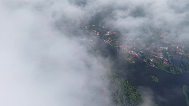 Aerial view of a foggy lake surrounded by dense green forest and scattered houses with red roofs, creating a calm, misty, and peaceful natural landscape at Tasik Raban