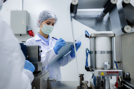 Female quality control inspector holding clipboard checking automated manufacturing machinery. Concept of industrial safety standard regulations in food or pharmaceutical plant, quality control.