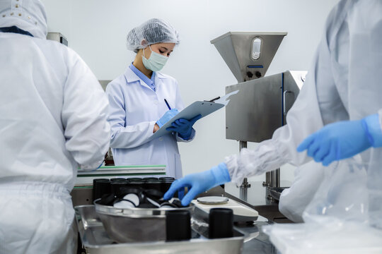 Factory laboratory inspector writing report on clipboard near automated capsule medicine packaging machine. concept of pharmaceutical packaging line, professional biotechnology manufacturing workflow.