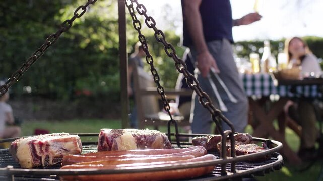 Steak and bratwurst sizzling on a swinging grill grate at a sunny garden barbecue with people in the background