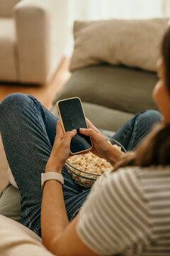 Woman relaxing on sofa using smartphone and eating popcorn