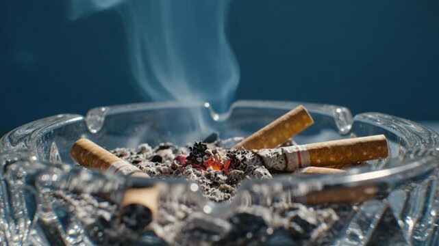 Close up of a glass ashtray with smoldering cigarette butts emitting wisps of smoke against a blue background