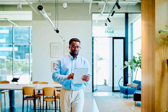 Professional African American businessman walking through a modern, bright office, actively working on a digital tablet
