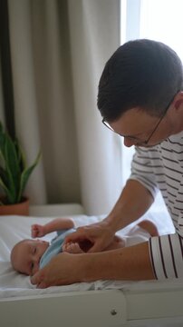 Young, attentive father with glasses carefully puts a light blue bodysuit on his adorable newborn son, who is lying on a white changing table in a brightly lit room at home