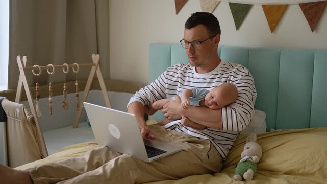 Young busy father attempting to work remotely on a laptop while holding his sleeping newborn baby in a cozy bedroom, illustrating the difficulties of balancing paternity leave and career