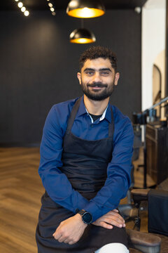 Adult African barber wearing dark-blue shirt and apron sitting in barber chair at barbershop