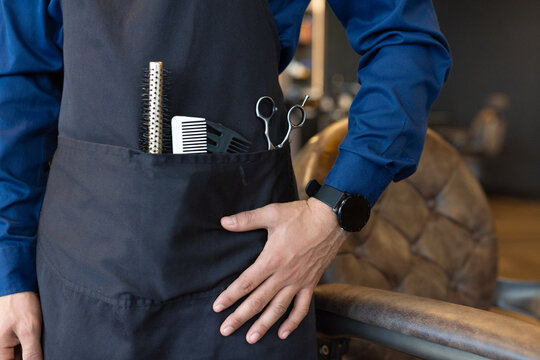 African American man wearing black apron holding round brush, combs, scissors at barbershop