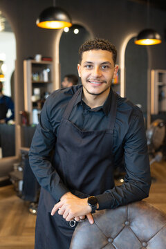 African male barbers leaning on tufted chair at modern barbershop with black apron holding scissors