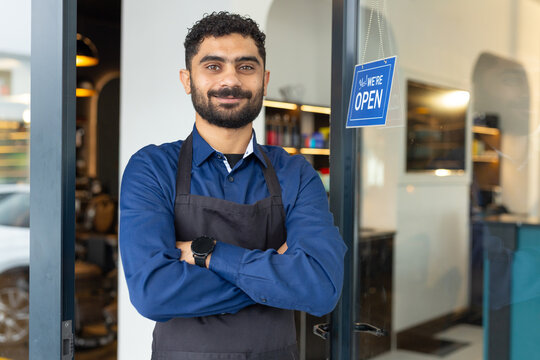 Adult male standing in shop doorway, wearing black apron and blue shirt, showing OPEN sign