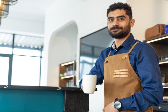 African American man standing behind cafe counter holding white mug wearing brown leather apron