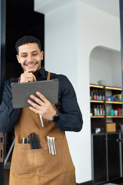 African male barber standing in shop doorway holding tablet, wearing brown apron with scissors