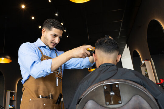 African American male barber and client trimming hair in barbershop with clippers and leather apron