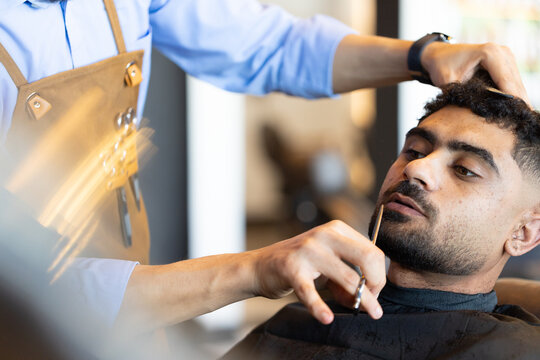 Diverse barber trimming African male client beard with trimmer in salon brown apron, copy space