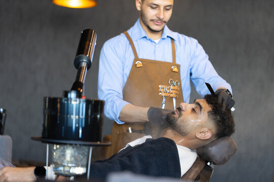 African American male barber trimming male client's hair at barbershop with apron and using clipper