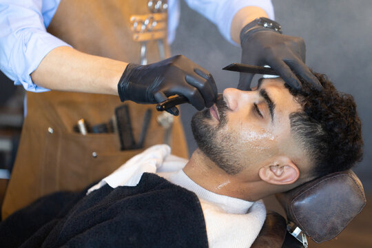 Barber in leather apron grooming African male client with straight razor and lather at barbershop