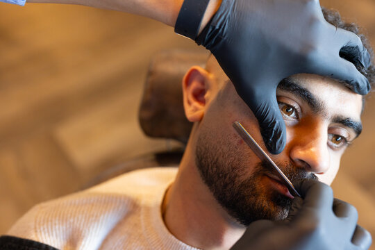 Adult male client sitting at barbershop getting beard shaping with straight razor and black gloves
