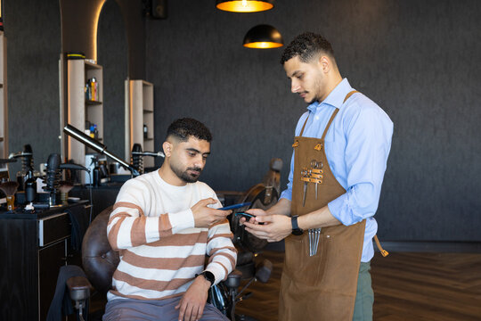 African American men barber and client comparing phones while seated in shop apron showing scissors