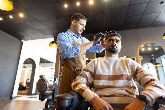 Male barber standing behind male client in barber chair cutting hair with clipper in barbershop