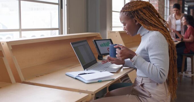 African American woman spotting tablet chart sipping mug typing on laptop noting data at window bar