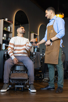African American male barber showing device to male client in barber shop, apron with scissors