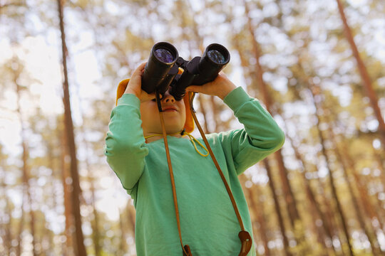 A child stands in a forest, using binoculars to observe nature. Trees surround the child in this outdoor setting. Sunlight filters through the branches